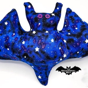 May include: A plush bat toy with a blue galaxy print, white stars, and purple accents. The bat has two purple button eyes and a whimsical design. The wings are spread wide, and the overall shape is soft and inviting.