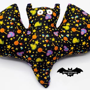 May include: A plush bat-shaped pillow with a black background covered in yellow stars, candy corn, and lollipops. The bat has button eyes and small ears. The Occasional Bat logo is in the lower right corner.