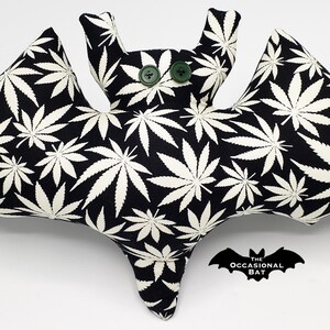 May include: A plush bat-shaped pillow with a black background and a white cannabis leaf pattern. The bat has green button eyes and a small logo that says "The Occasional Bat".