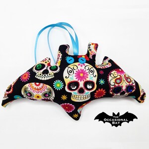 May include: A black bat-shaped ornament with a blue ribbon hanger. The bat is covered in a colorful fabric with a Day of the Dead sugar skull pattern. The fabric features white, pink, yellow, green, and blue colors.