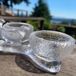 May include: A clear glass bird-shaped candle holder with two bowls. The candle holder is made of textured glass and sits on a wooden surface. The background is blurred, showing a view of the water and sky.