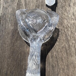 May include: Clear glass bottle stopper with a floral design. The stopper has a textured stem and a rounded top with a flower-like shape. The glass is transparent, allowing light to pass through.