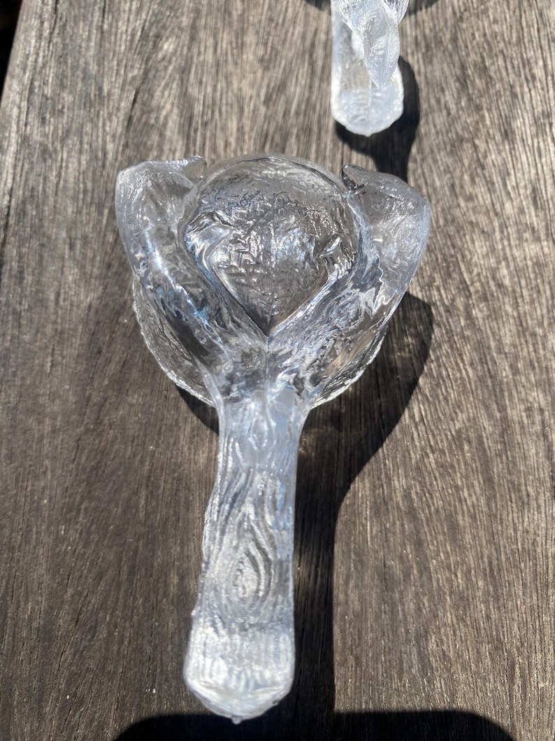 May include: Clear glass bottle stopper with a floral design. The stopper has a textured stem and a rounded top with a flower-like shape. The glass is transparent, allowing light to pass through.