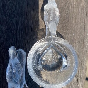 May include: Two clear glass bird-shaped bowls. Each bowl features a bird perched above a round, textured bowl. The glass has a textured, frosted appearance. The bowls are sitting on a wooden surface.