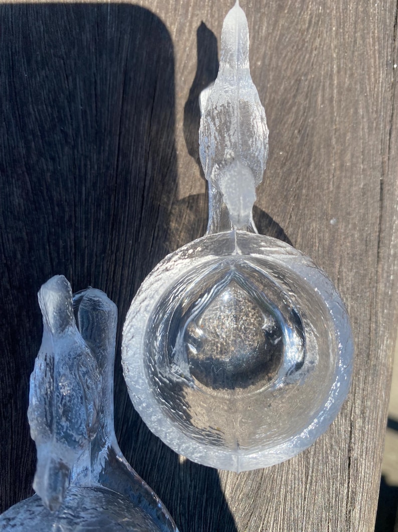 May include: Two clear glass bird-shaped bowls. Each bowl features a bird perched above a round, textured bowl. The glass has a textured, frosted appearance. The bowls are sitting on a wooden surface.
