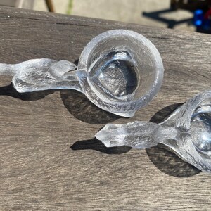 May include: Two clear glass measuring cups with bird-shaped handles. The cups are made of textured glass and sit on a wooden surface. The cups are empty and appear to be new.