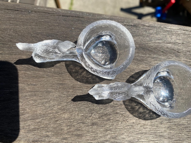 May include: Two clear glass measuring cups with bird-shaped handles. The cups are made of textured glass and sit on a wooden surface. The cups are empty and appear to be new.