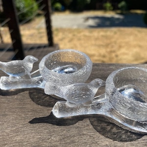 May include: Clear glass candle holders with a textured surface. Each holder features a bird perched on a branch. The candle holders are designed to hold small candles and add a decorative touch to any space.