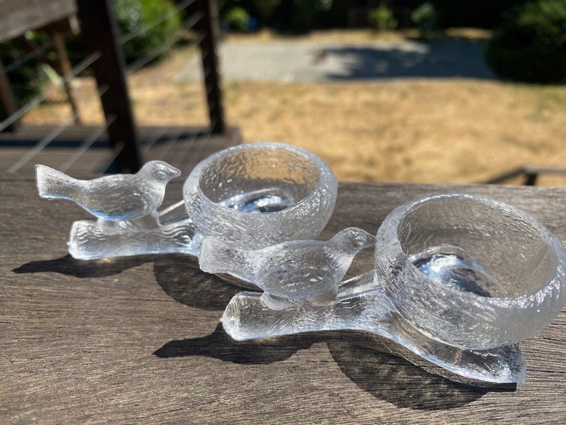 May include: Clear glass candle holders with a textured surface. Each holder features a bird perched on a branch. The candle holders are designed to hold small candles and add a decorative touch to any space.