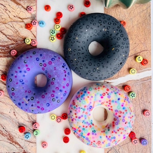 Op de afbeelding: Drie kleurrijke donuts met sprinkles. De donuts zijn zwart, paars en wit met kleurrijke sprinkles. De donuts liggen op een wit oppervlak met kleurrijke kralen.