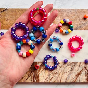 Può includere: Una collezione di anelli di perline colorate, tra cui viola, rosa, blu e arcobaleno. Gli anelli sono fatti di piccole perline rotonde e sono disposti su una superficie di legno.