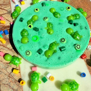 May include: A bright green slime, circular in shape, adorned with small green turtle figures and decorative beads. The slime is surrounded by colourful beads and shapes. The turtles are scattered throughout the slime.