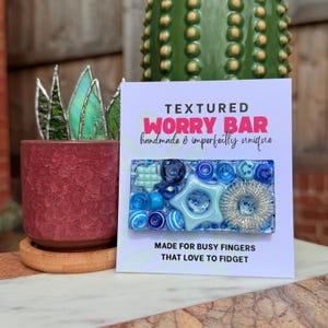May include: A rectangular worry bar featuring various blue and light blue tactile elements, including stars, beads, and a silver-toned coil, mounted on a white display card with the text "TEXTURED WORRY BAR" and "MADE FOR BUSY FINGERS THAT LOVE TO FIDGET."