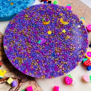 Op de afbeelding: Een paarse en gouden harsonderzetter met een maan- en sterrendesign. De onderzetter is gevuld met kleine paarse en gouden kralen.