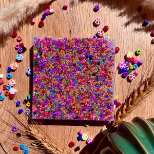 May include: A square resin coaster with a colourful mix of small beads embedded in the surface. The coaster is surrounded by colourful beads and other decorative items.
