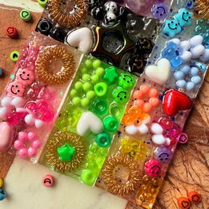 Puede incluir: Una colección de bloques rectangulares de resina incrustados con una variedad de cuentas de plástico coloridas, incluyendo caras sonrientes, corazones, estrellas, flores y espirales doradas, dispuestos en un vibrante patrón de mosaico.