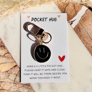 Könnte beinhalten: Ein schwarzer und goldener Schlüsselanhänger "Pocket Hug" mit einem Smiley-Anhänger. Die Karte lautet "Hier ist ein kleiner Taschen-Umarmung, bitte bewahre ihn sicher und nah auf. Dann wird er da sein, neben dir, wenn du ihn am meisten brauchst."