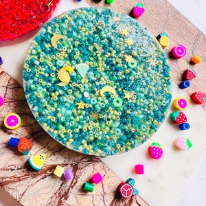May include: A circular resin coaster filled with teal, green, and yellow beads, decorated with gold stars and crescent moons. The coaster is surrounded by colourful fruit-shaped beads, such as strawberries, lemons, and watermelons. A red, textured resin piece is also visible.