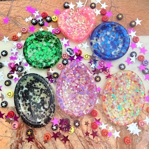 May include: Five resin trinket dishes in heart, oval, and circular shapes, each filled with colorful star and moon glitter. The collection includes pink, green, blue, black, and iridescent clear pieces, surrounded by loose star confetti and beads.