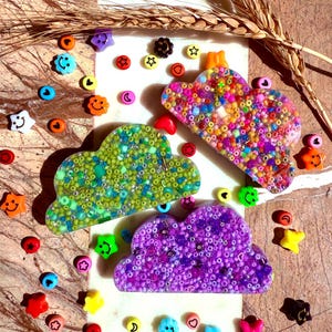 Op de afbeelding: Drie wolkvormige ornamenten van hars, gevuld met gekleurde kralen. De wolken zijn groen, paars en veelkleurig. De wolken zijn omringd door kleine gekleurde kralen.