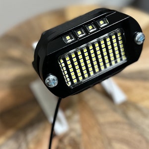 May include: A black LED light with a rectangular array of white LEDs. The light is mounted on a black bracket with two screws.