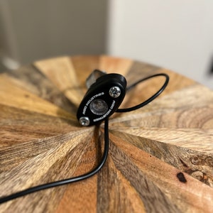 May include: A black LED light with the text "RIGID INDUSTRIES" and two silver screws. The light has a clear lens and a black wire extending from it. The light is on a wooden surface.