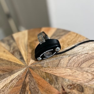 May include: A black round LED light with a silver clamp. The light has the text "Rigid Industries" printed on it.