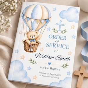 May include: A white booklet titled ORDER OF SERVICE Of William Smith For His Baptism, Saturday, 23 August 2026, St Augustine's Church. The cover features a watercolor illustration of a teddy bear in a blue and tan hot air balloon with clouds, stars, and a blue cross.