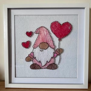 May include: String art picture of a gnome holding a red heart balloon, with two small red hearts. The gnome has a pink hat and outfit, a white beard and brown face. The artwork is in a white frame.