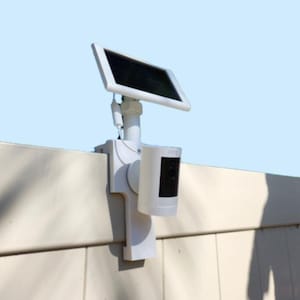 May include: A white security camera mounted on a fence with a white solar panel attached to the top. The camera is cylindrical with a black front face and the word Ring printed on the side. A white cable connects the solar panel to the camera unit.