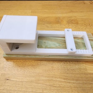 May include: White plastic 3D printed jig with a wooden base. The jig has a rectangular opening and two screw holes.