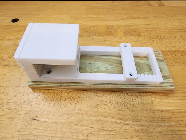 May include: White plastic 3D printed jig with a wooden base. The jig has a rectangular opening and two screw holes.
