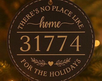 May include: A round, black Christmas ornament with gold text that reads "THERE'S NO PLACE LIKE home 31774 FOR THE HOLIDAYS." The ornament is hanging from a gold string. A red ornament is in the background.