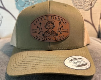 May include: An olive green and tan trucker hat with a curved brim. The hat features a leather patch with the text "Little Debbie Hunting Team" and a graphic of a woman with bows. The hat has a snapback closure.