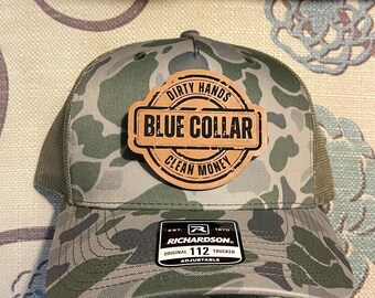 May include: Camouflage pattern trucker hat with a brown leather patch that reads "Dirty Hands Blue Collar Clean Money." The hat is adjustable and features a Richardson 112 Trucker label.