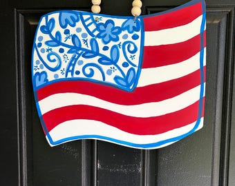 May include: A wooden American flag-shaped door hanger. The flag features red and white stripes with a blue corner adorned with blue floral designs. A string of wooden beads forms the handle for hanging.