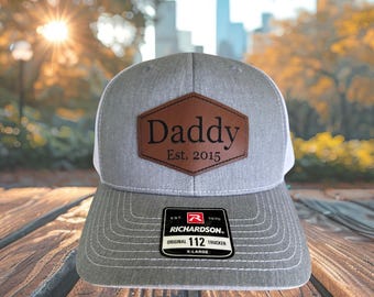 May include: A gray and white trucker hat with a brown leather patch that reads "Daddy Est. 2015." The hat has a curved brim and a mesh back. The Richardson 112 Trucker hat is X-Large.