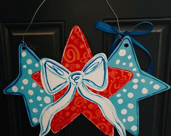 May include: A patriotic star-shaped door hanger with a red star in the center, adorned with a white bow and blue accents. The outer stars are blue with white polka dots. A blue ribbon is attached for hanging.