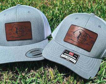 May include: Two gray trucker hats with white mesh backs. Each hat has a brown leather patch with a fist bump design and the word "DAD" and initials "JB". One hat has a "Richardson 112" tag.