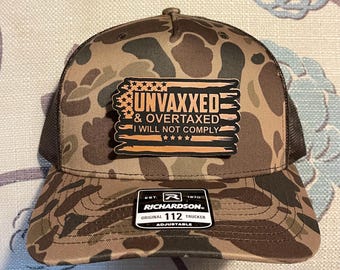 May include: Camouflage trucker hat with a brown mesh back and a patch that reads "UNVAXXED & OVERTAXED I WILL NOT COMPLY." The hat is adjustable and features the Richardson brand logo.