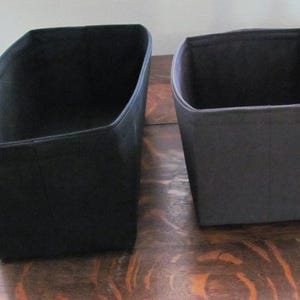 May include: Two black fabric storage bins of different sizes. The bins have a rectangular shape with slightly angled sides. The bins are sitting on a wooden surface.