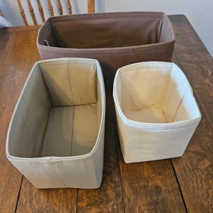 May include: Three fabric storage bins in varying sizes and colors. The largest bin is a dark brown, rectangular shape. Two smaller bins are a light beige color, one rectangular and one square. All bins are empty.