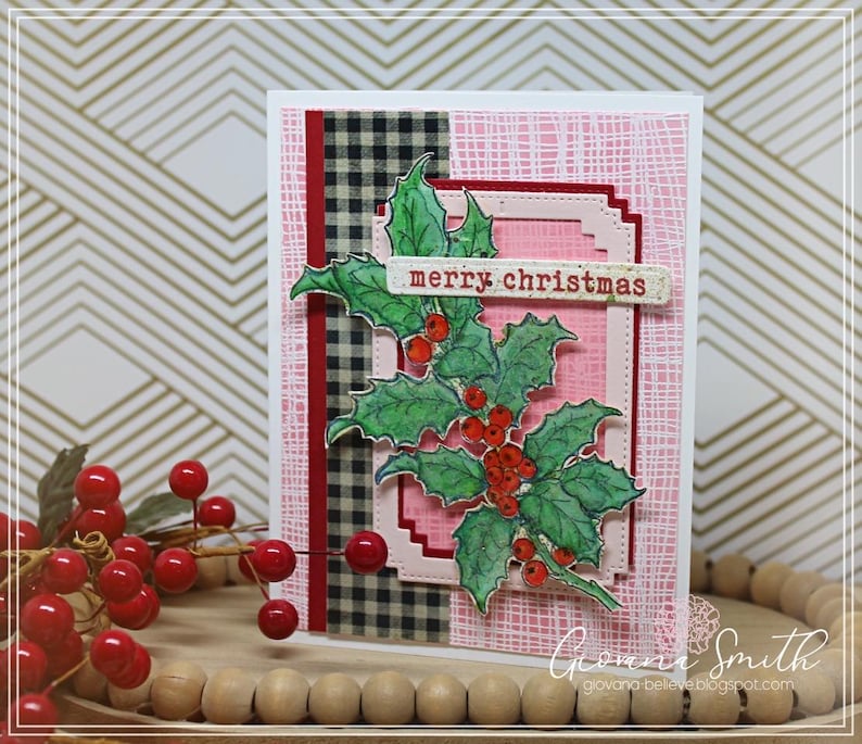 May include: A handmade Christmas card featuring a holly branch with red berries and the text "merry christmas." The card has a pink background with a plaid and textured design. The card is on a wooden tray with red berries.