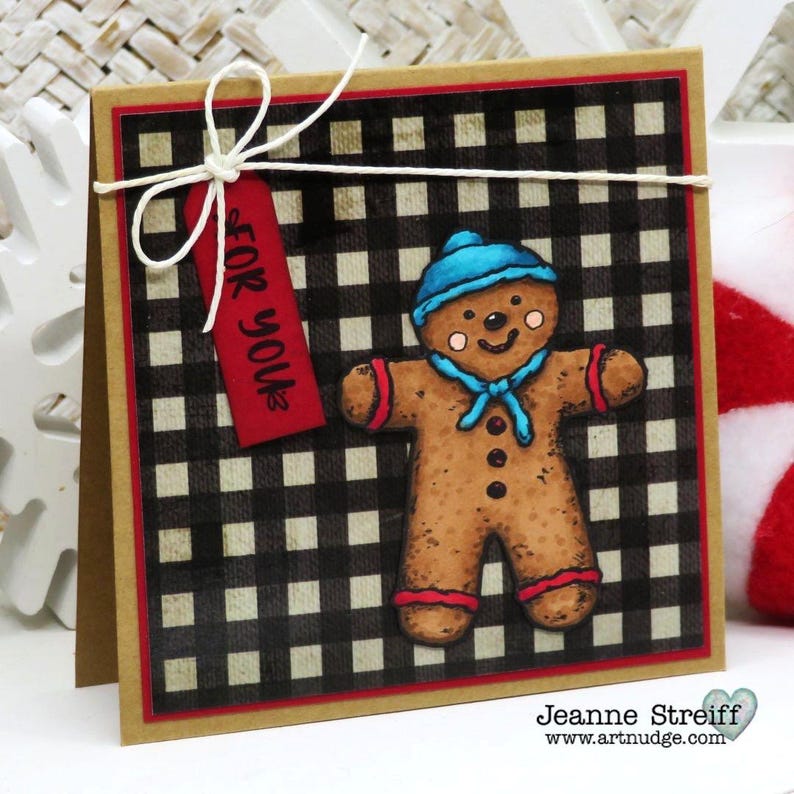 May include: A handmade Christmas card featuring a gingerbread man with a blue hat and scarf on a black and white checkered background. A red tag with "FOR YOU" is tied with white string.