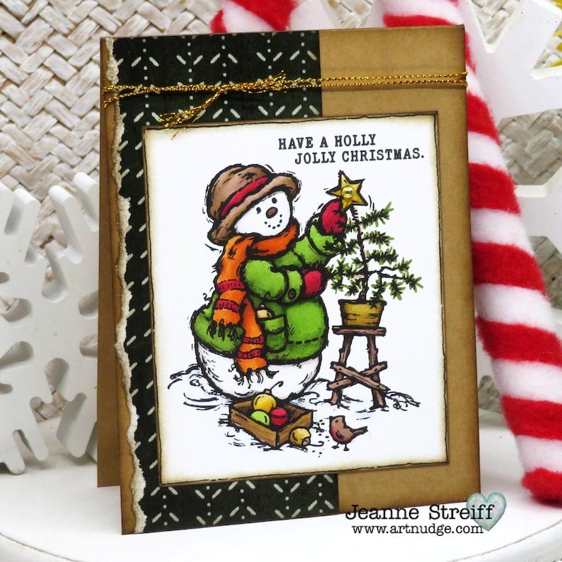 May include: A Christmas card featuring a snowman decorating a small tree. The snowman wears a brown hat, orange scarf, and green coat. The card has the text "Have a Holly Jolly Christmas." A small bird and a box of ornaments are also shown.