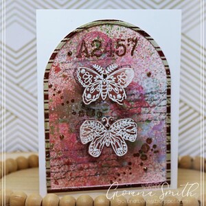 May include: A handmade greeting card featuring a decorative arch design with a textured, multi-colored background. Two white butterfly cutouts are layered over the design. The numbers "A2 57" are at the top. The artist's signature is at the bottom.