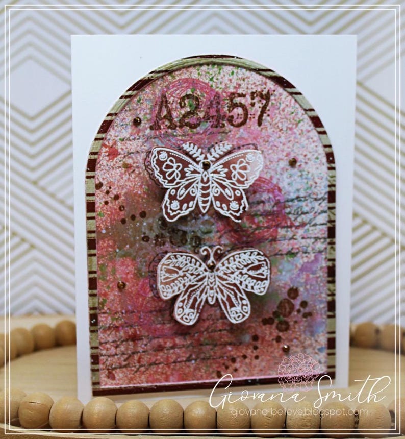May include: A handmade greeting card featuring a decorative arch design with a textured, multi-colored background. Two white butterfly cutouts are layered over the design. The numbers "A2 57" are at the top. The artist's signature is at the bottom.