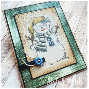May include: A handmade Christmas card featuring a snowman with a green and white striped scarf, a blue hat, and a blue button. The card has a green and brown border with a white polka dot pattern.