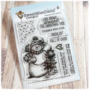 May include: A set of clear photopolymer stamps by SweetMissDaisy Designs featuring a snowman, a small pine tree, a heart, and holiday-themed text including Love Grows Around The Christmas Tree, Trimmed with Love, and Fresh Cut and Full of Cheer.