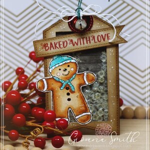 May include: A handmade Christmas tag featuring a gingerbread man with a blue hat and scarf. The tag reads "BAKED WITH LOVE" and includes a small bell and red berries. The tag is brown with a clear window.